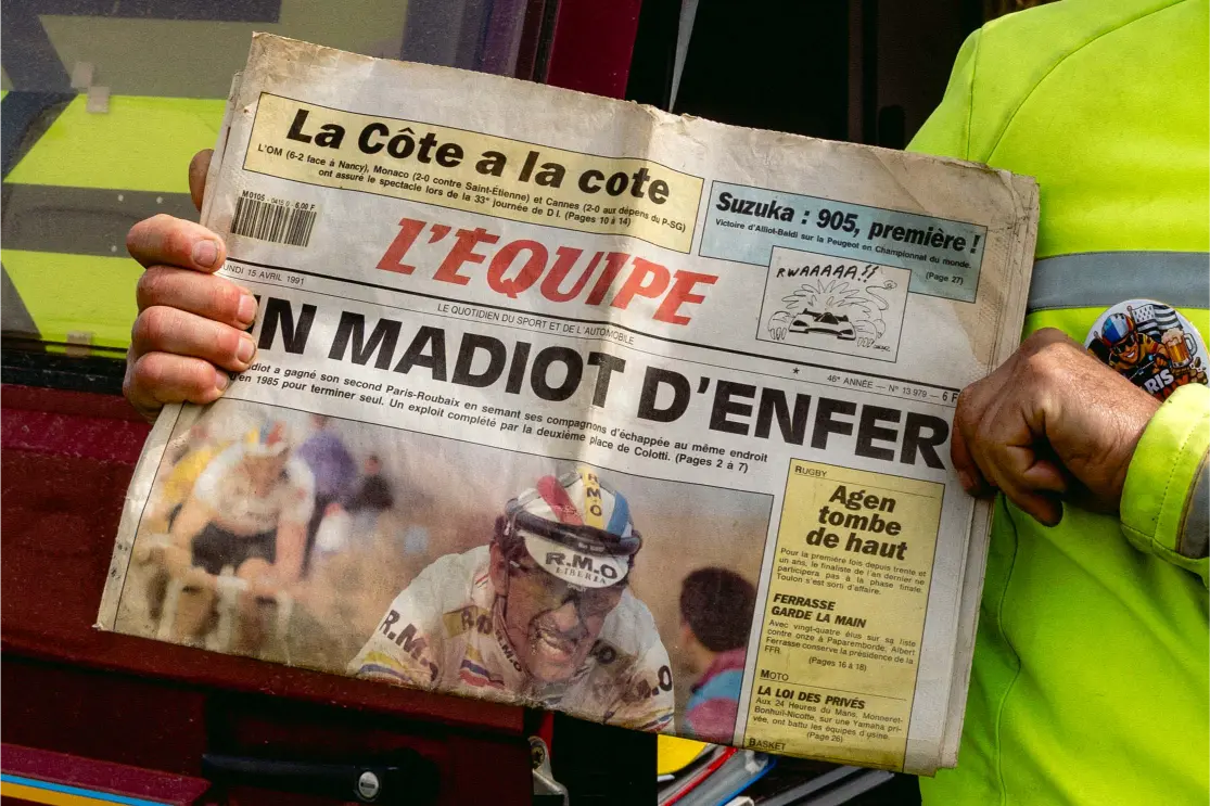 An older man holding up an original L'Equipe newspaper from 1991 by the course
