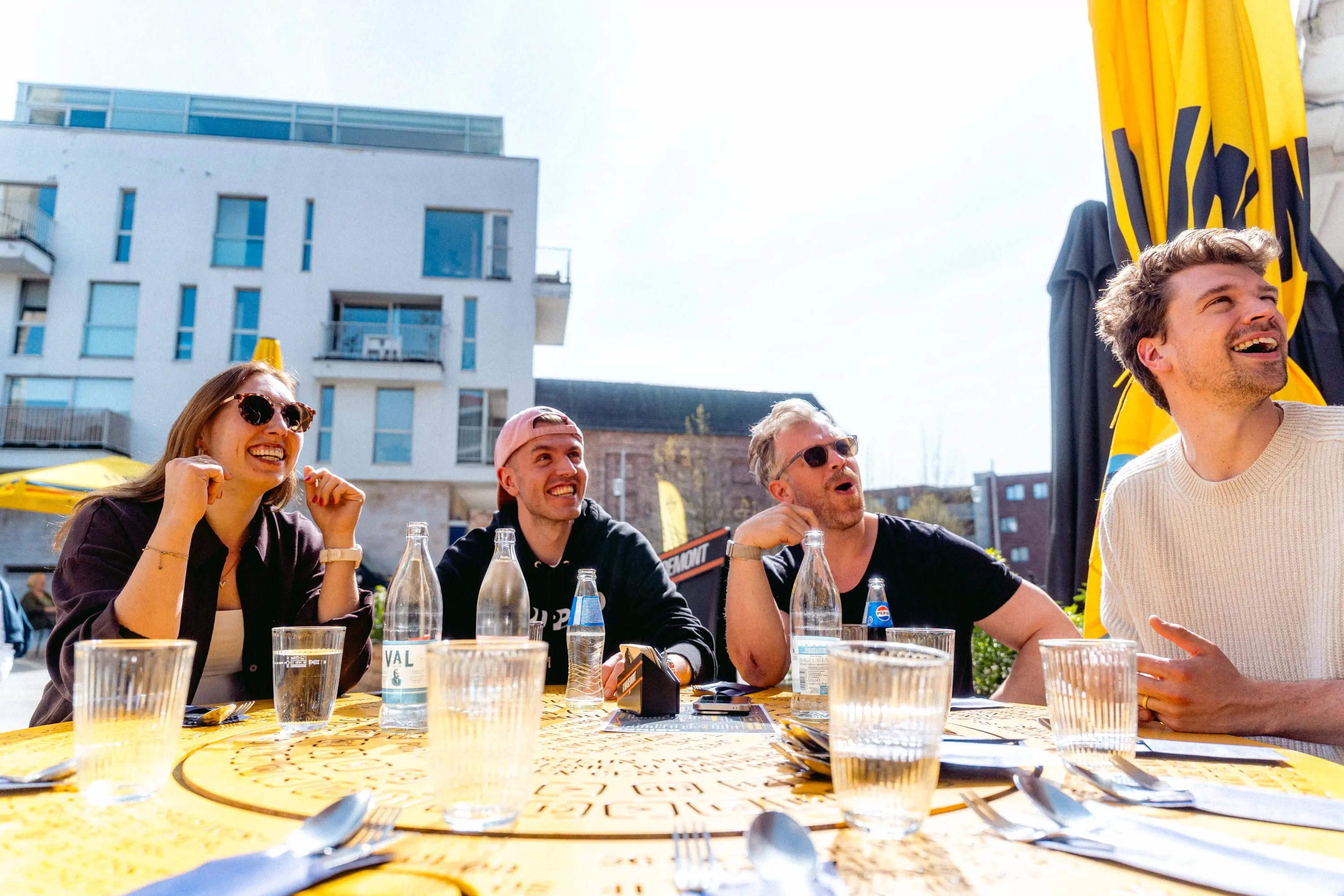 Reaktion auf den Trikot-Drop: vier lachende Gesichter am Café-Tisch unter dem gelben Van-Rysel-Schirm in Lille