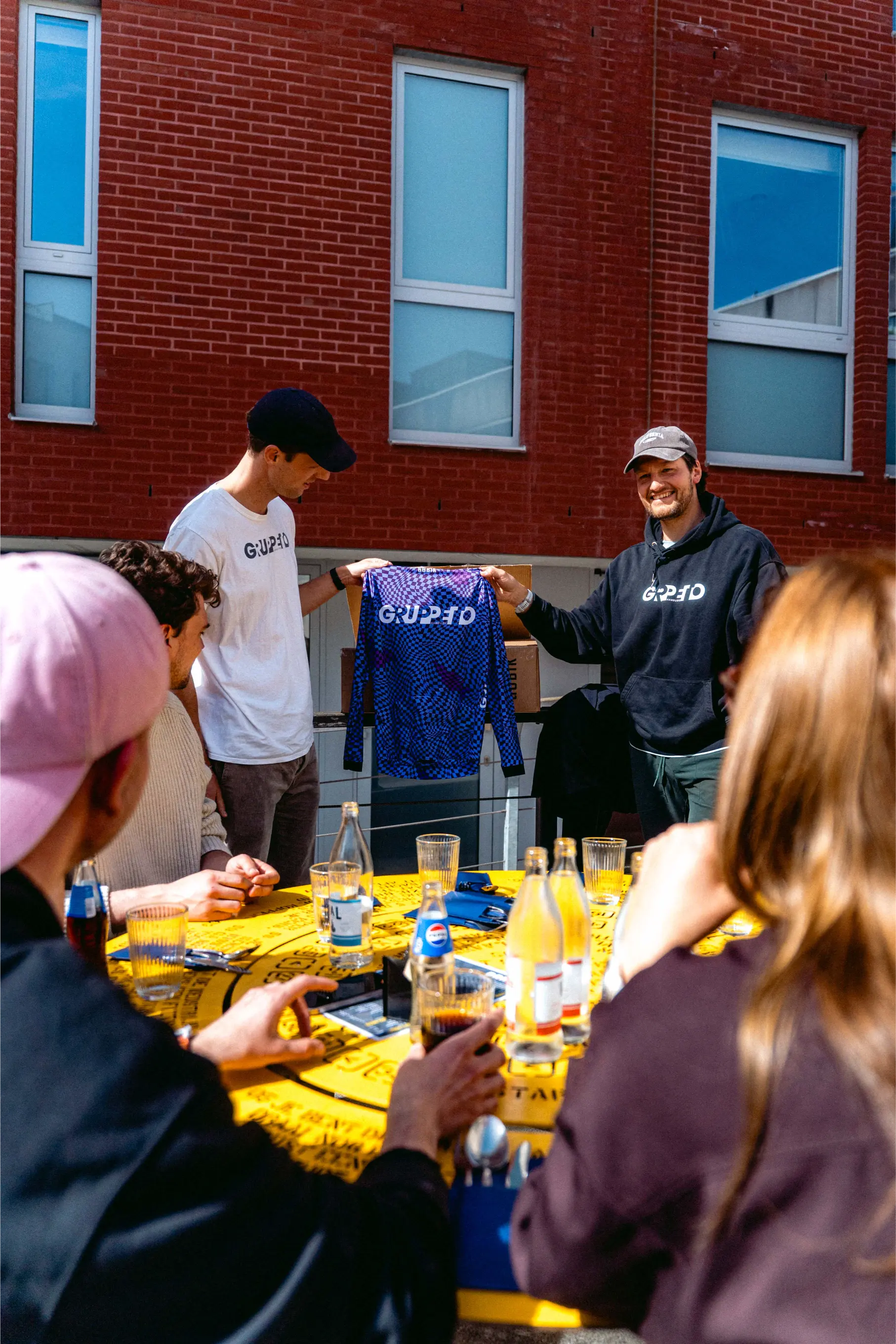 Jemand hält das neue violett-schwarze Road-Captains-Trikot von Gruppetto × Van Rysel vor einem gelben Tisch hoch